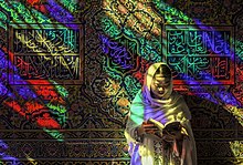 Framför en vägg fylld med skrifter lutar en kvinna upplyst av glasmålningsfönstret ansiktet mot en bok som hon håller platt i händerna.