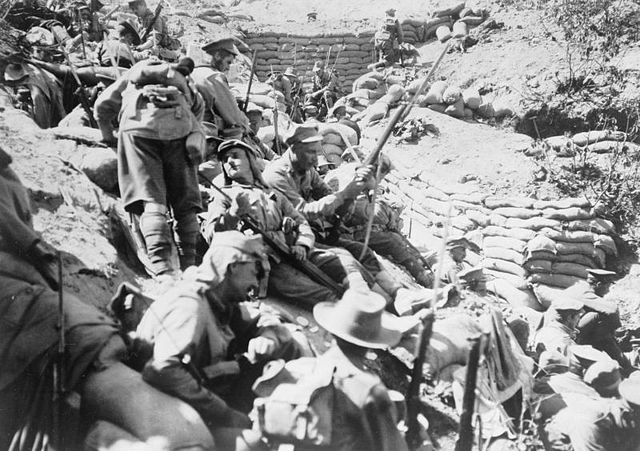 Soldiers with rifles, slouch hats and service caps sit on a steep escarpment behind a sandbagged trench