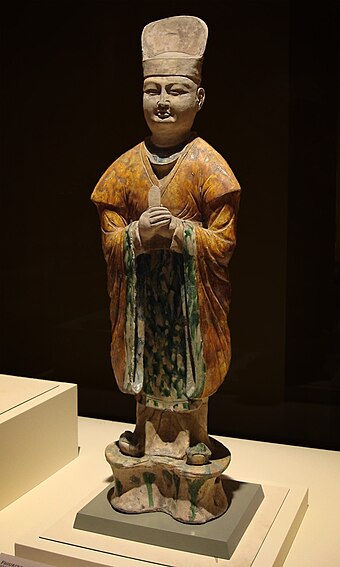 Tang tomb figure of an official dressed in Hanfu, with a tall hat, wide-sleeved belted outer garment, and rectangular "kerchief" in front. A white inner gown hangs over his square shoes. He holds a tablet to his chest, a report to his superiors.