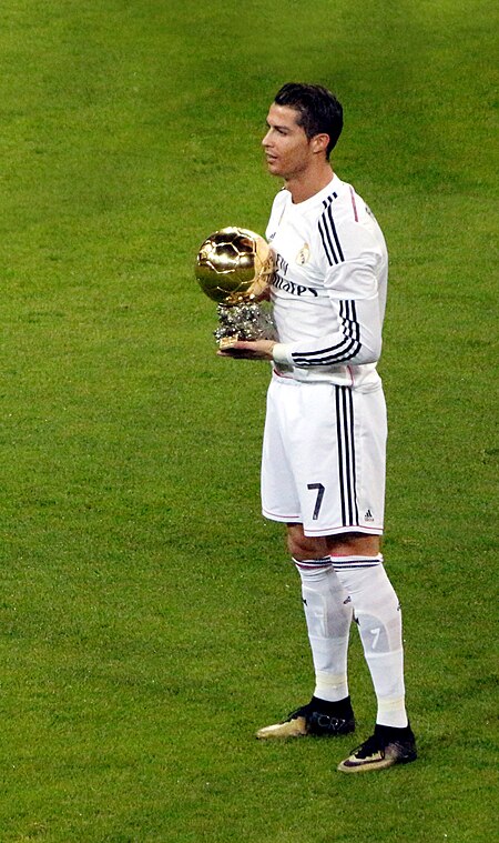 A man in a white shirt poses with a golden ball while standing on football pitch.