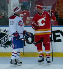Photographie de deux joueurs de hockey sur glace discutant. L'un porte un uniforme blanc, bleu et rouge, l'autre rouge, blanc et jaune.
