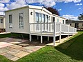 Image shows a cream static caravan surround by grass and a patio with ramp up to entrance.