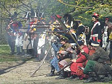 Grupa rekonstruktorów historycznych w mundurach polskiej XVIII-wiecznej piechoty oraz w strojach cywilnych z epoki oddaje salwę.