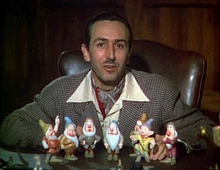 A color photograph of Walt Disney sitting behind a desk. Seven figurines are standing before him.