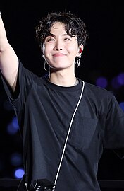 J-Hope sonriendo y saludando con la mano izquierda.