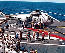 Photographie en couleur de l'équipage d'Apollo 16 quittant l'hélicoptère de récupération Sikorsky SH-3 Sea King, posé sur le porte-avions USS Ticonderoga. Ils sont pris en photo par de nombreuses personnes présentes sur la piste du porte-avions.