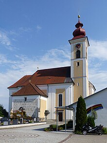 Die Pfarrkirche bei Sonnenschein und blauem Himmel fotografiert. Rechts im Bild der Kirchturm mit rot lackiertem Zwiebelhelm, dem sich links das niedrigere Langhaus mit Chor und dem Sakristeianbau anschließt. Vor der Kirche ist ein Stück des umgebenden Friedhofs zu sehen.