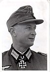 A man wearing a military uniform and field cap with an Iron Cross displayed at the front of his uniform collar.