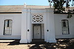 Just a short distance higher up from La Gratitude on the left side of Dorp Street, stands a modest little building with a peculiar verandah. It looks rather out of place among the rest of the buildings by which it is squeezed in. This is the Stellenbosch Gymnasium from which the Paul Roos Gymnasium and the University of Stellenbosch developed. Gimnasia 120 Dorp Street Stellenbosch.JPG