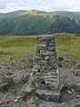 The trig point at the summit of Loughrigg, with Fairfield in the background.