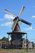 Photographie en couleurs d'un moulin à vent, d'une habitation et d'un entrepôt entouré d'une voie urbaine et de bâtiments.