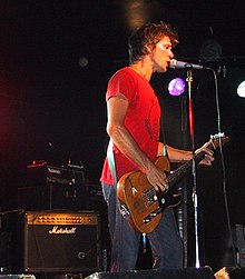 Um homem é mostrado em perfil lateral de três quartos.  Ele está cantando em um microfone enquanto toca um violão.  Ele usa uma camiseta vermelha e jeans.  Atrás dele estão as luzes do palco e um alto-falante amplificado.