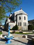 Photo représentant le chevet polygonal ensoleillé de l'église Saint-Jean-Saint-Charles surmonté d'une statue. À l'arrière, on peut voir une partie du clocher carré. Au premier plan sur la gauche se trouve une fontaine métallique de couleur bleu turquoise avec vasque.