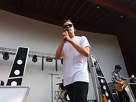 Petr Lexa wearing a white t-shirt, dark pants, and dark sunglasses, holding a microphone onstage, with band in the background