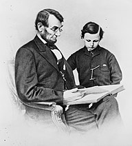 A seated Lincoln holding a book as his young son looks at it