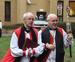 Justin Welby och Stephen Cottrell i vit röcklin, svart chimere och röd akademisk hood. Cottrell håller en biskopskräkla i silver i handen.