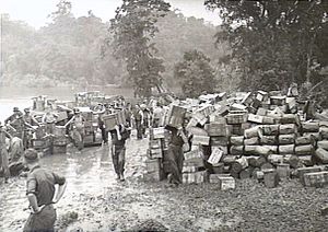 Schwarz-Weiß-Foto einer großen Anzahl kleiner Holzkisten, die auf schlammigem Boden gestapelt sind, wobei Männer in Militäruniformen zusätzliche kleine Holzkisten tragen