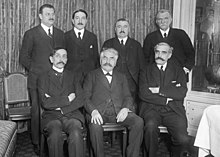 Photo en noir et blanc de sept hommes moustachus portant un costume (trois assis, au premier plan, et quatre debout, à l’arrière-plan)