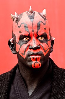 Homme dont le visage est peint en rouge et possède des tatouages faciaux noirs.