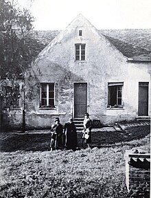 La dernière Solitaire de Port-Royal, vêtue d'une longue robe noire, coiffée d'un foulard, tenant un bâton comme canne, entre deux visiteuses devant son humble maison à laquelle on accède par un escalier de quatre marches et comportant deux pièces au rez-de-chaussée et deux autres à l'étage en soupente, l'une avec fenêtre en façade et l'autre avec fenêtre ouvrant sur l'étang des moniales à l'arrière de la maison au-dessus du talus.