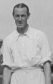 A black-haired man in a white shirt with sleeves rolled up looks into the camera, posing