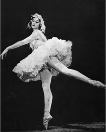 Photographie d'une ballerine dans un tutu court et une coiffe à plumes, debout en pointe sur sa jambe droite avec son bras gauche et sa jambe gauche étendus à des angles de 45 degrés par rapport à la ligne verticale de sa jambe droite et de son torse.