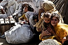 Line of women wearing hijabs sitting on the ground. Some carry bags or children.