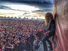 A man with long, curly hair standing on a stage with an open mouth while playing bass guitar. He points his bass guitar toward the photographer and bends his knees. In the background, the sun sets on the festival-like fairground behind the audience.