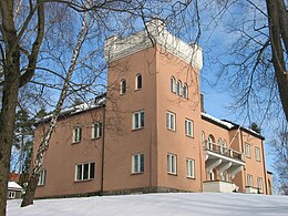 Un edificio importante en piedra rosada o revestimiento, formado por una torre de tres pisos con alas que tienen dos. El edificio se alza, enmarcado por árboles, en un jardín cubierto de nieve.