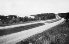 A black and white photograph shows two parallel gravel roads curving through an artificial clearing of an otherwise forested area