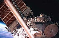 Um homem vestido com um traje espacial visto rastejando ao longo de um módulo de estação espacial cilíndrico branco.  Um grande painel solar pode ser visto projetando-se do topo do módulo e várias outras peças do aparelho são visíveis.  O horizonte e o espaço da Terra são visíveis por trás do painel solar.