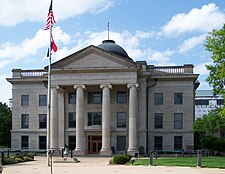 A light-colored three-story stone building with an American flag flying from a pole in front above another red, white and blue striped flag. The building has a pedimented front pavilion with four columns and a small dome at the top from which another American flag is being flown.