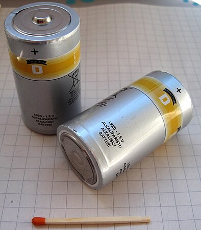 Two D cell batteries, similar to those used in the Electro Tic-Tac-Toe. These large alkaline batteries were standardized and could commonly be found in stores. These batteries were not typically integral in devices, and could be changed on the fly, but this was because these batteries were one use only and not meant to be rechargeable. A match stick is included for scale.