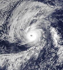 A photograph of a powerful hurricane over the Eastern Pacific Ocean. It has a clear eye surrounded by a small but nearly axisymmetrical area of deep convection. A large area of high clouds is fanning out to the north and west of the hurricane, with a pronounced band spiraling out to the south and southwest. A small and thin area of convection, oriented north to south, is to the right of Kenneth.