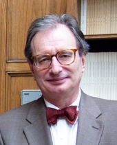 Photographie d'un homme portant une veste de costume grise, un nœud papillon rouge et des lunettes.