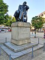 gleiche Fotoperspektive, Skulptur ist schwarz, Inschrift nun in weiß, im Hintergrund blühende Bäume und parkende Autos.