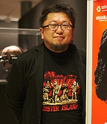 Higuchi stands next to a poster of his 2016 film Shin Godzilla. He wears a T-Shirt emblazoned with the text "Greetings from Monster Island", featuring an illustration of Megalon, Gigan, Godzilla, and Jet Jaguar posing in the 1973 film Godzilla vs. Megalon.