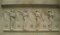 Das Hauptportal der katholischen de:St. Fideliskirche (Stuttgart) in Stuttgart wird von zwei Hochreliefs mit je vier musizierenden Engeln flankiert. Die zum Portal gewandten Engel sind mit großen, weit ausladenden Flügeln dargestellt, tragen fußlange Gewänder und ihr Haupt ist verhüllt oder zeigt einen lockigen Haarschopf. Sie „schweben“ mit den Zehenspitzen ihrer nackten Füße auf Kugeln, die auf einer Zickzackleiste ruhen. Das Traufgesims über ihren Köpfen schützt sie vor Regen. Zwischen den Engeln sind Rundmedaillons angeordnet, die in einem doppelten achtzackigen Stern christliche Symbole bergen. Die Abbildung zeigt die linke Relieftafel. Attribute der Engel von links nach rechts: Buch, Tamburin, Laute, Lyra. Symbole von links nach rechts: Abendmahlskelch mit mit schwebender Hostie, Lateinisches Kreuz, Golgathakreuz, flammendes Herz Jesu, Buchstabe A. Die Buchstaben Alpha und Omega sind das Symbol für Christus als Anfang und Ende (Offenbarung des Johannes 22, 13). Die Symbolreihe der rechten Relieftafel beginnt mit dem Buchstaben Ω.