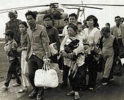 Twelve refugees of varying ages, carrying bundles of possessions, arrive on the deck of a United States naval vessel. Three US airmen, as well as a helicopter, are visible in the background.