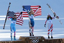 Foto av tre kvinnelige skiløpere på et podium, to av dem vinker USAs flagg, den andre bærer skiene.