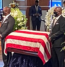 Two men wearing dark business suits stand on either side of a casket. The casket has been draped with a flag, a sign of military or government service in that country