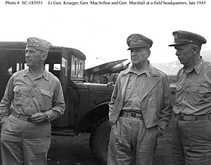 Three men in Army uniforms with open neck shirts. Krueger wears a garrison cap, MacArthur his special cap, and Marshall, a peaked cap. in the background there is a truck and a propeller driven airplane.