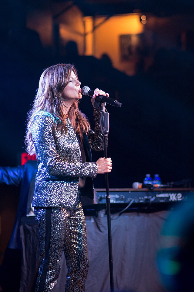 A woman with long brown hair wearing a silver jacket and pants, singing into a microphone