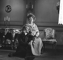 Photo noir et blanc.Homme en noir assis aux pieds d'une dame, un bras sur ses genoux, la regardant.