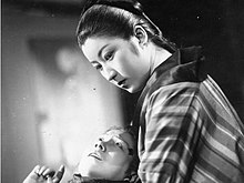 Hier ist eine Fotografie in schwarz-weiß und im Querformat zu sehen. Urabe Kumeko ist in der Bildmitte und hält eine liegende Person.