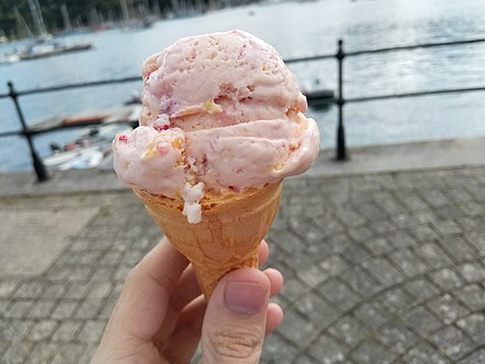 A single coned scoop of tutti frutti ice-cream, with a blurred background of a British harbour.