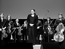 Photographie en noir et plan d’une femme debout, se tenant devant quelques musiciens.