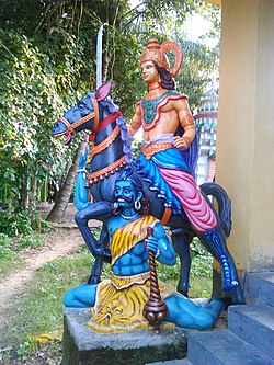 Kalki sits on a black horse with a sword on his hand, slaying a man, which could represent evil, who is laying on the floor with his legs spread out in an uncomfortable position. The evil man holds a club and wears tiger skin as clothing.