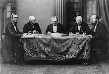 Photographie noir et blanc de cinq hommes en complet assis autour d'une table de réunion.