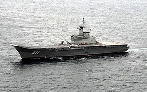 A small aircraft carrier with an upturned bow underway in calm seas. The number "911" is painted on the side, and an AV-8S aircraft and S-70 helicopter are sitting on the flight deck.